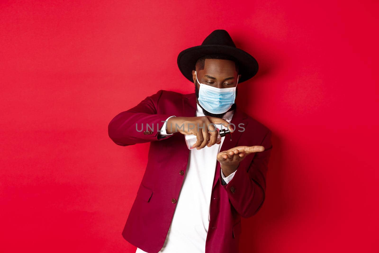 Covid-19, quarantine and holidays concept. Handsome Black man in face mask and party outfit, disinfecting hands with hand sanitizer, using antiseptic, standing over red background.