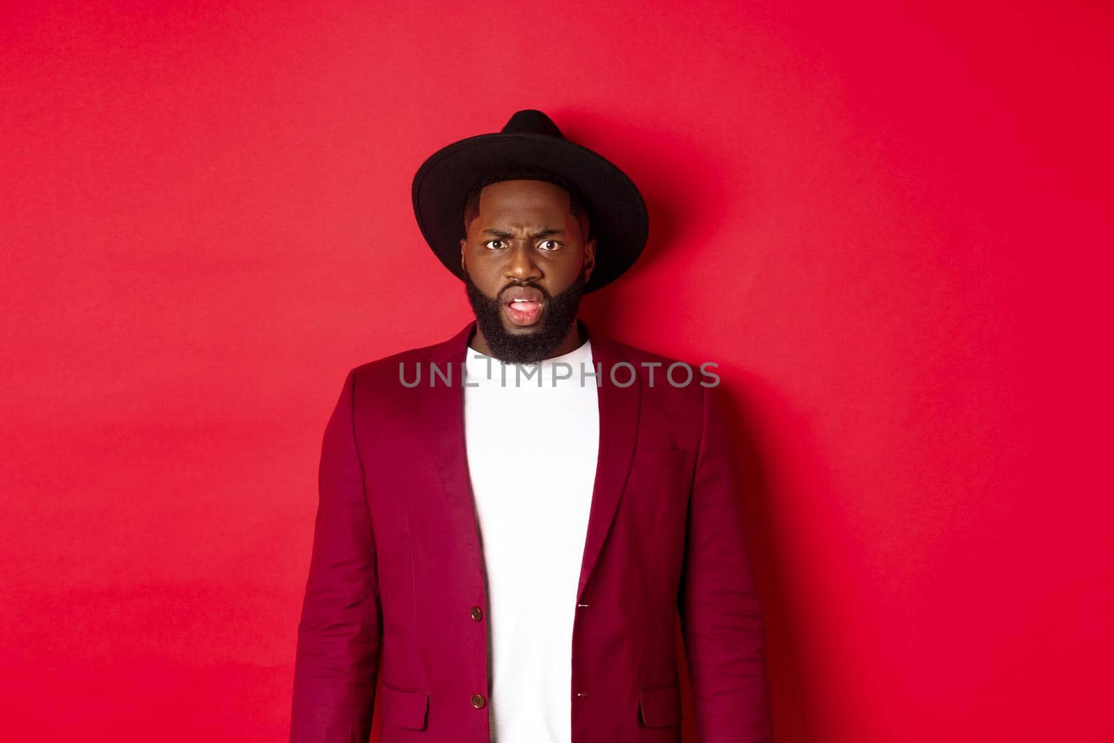 Fashion and party concept. Confused Black man in classy outfit frowning and staring at camera displeased, cant understand, standing over red background.