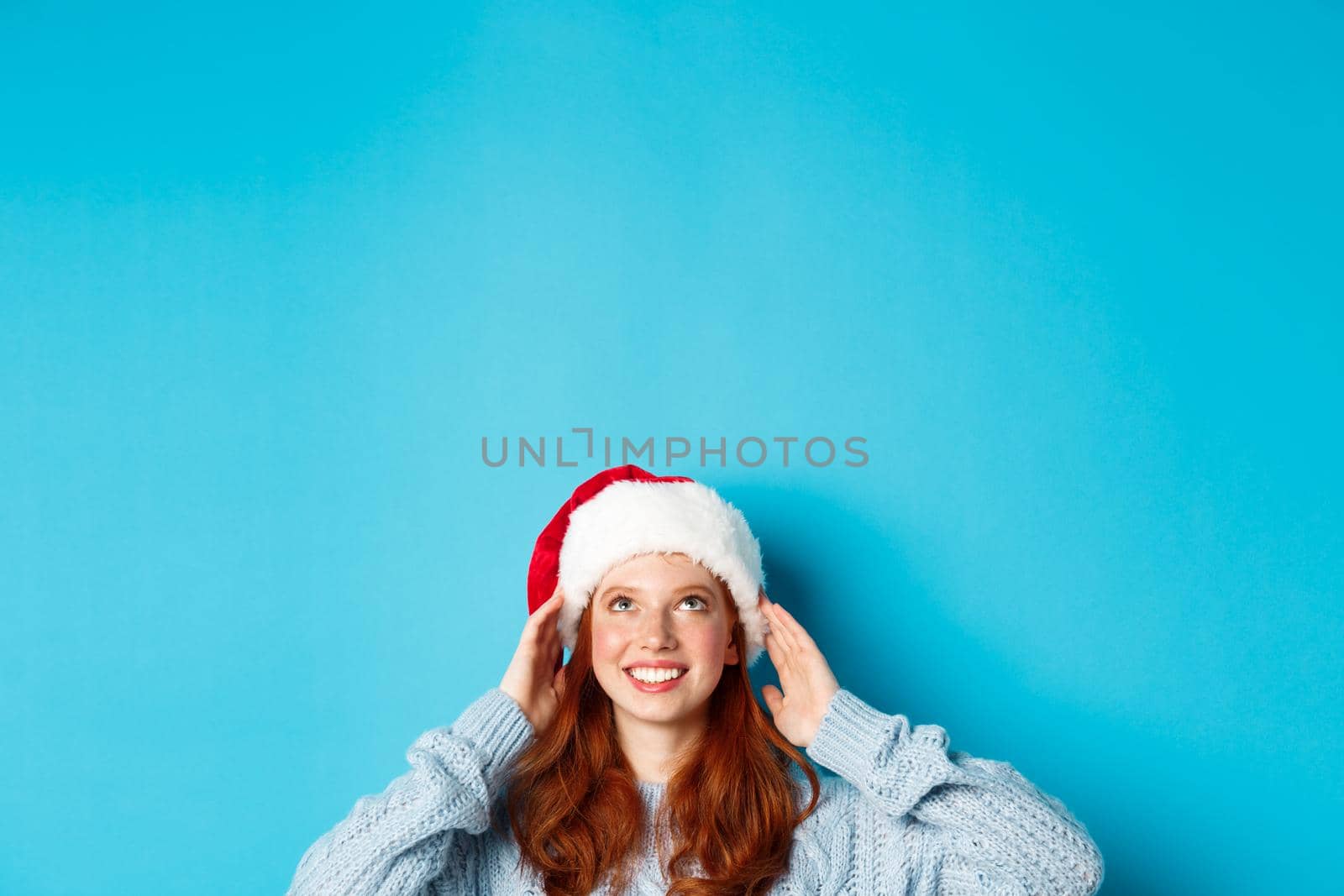 Winter holidays and Christmas eve concept. Head of cute redhead girl in santa hat, appear from bottom and looking up at copy space, staring logo, standing over blue background.