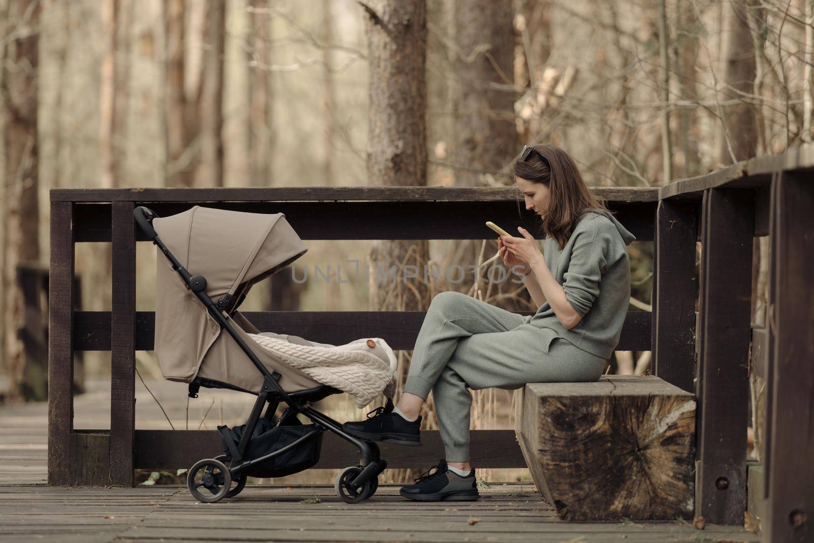 The young mom is working with her cellphone while her infant child is sleeping in the baby stroller. The mother with her child in the park for fresh air.