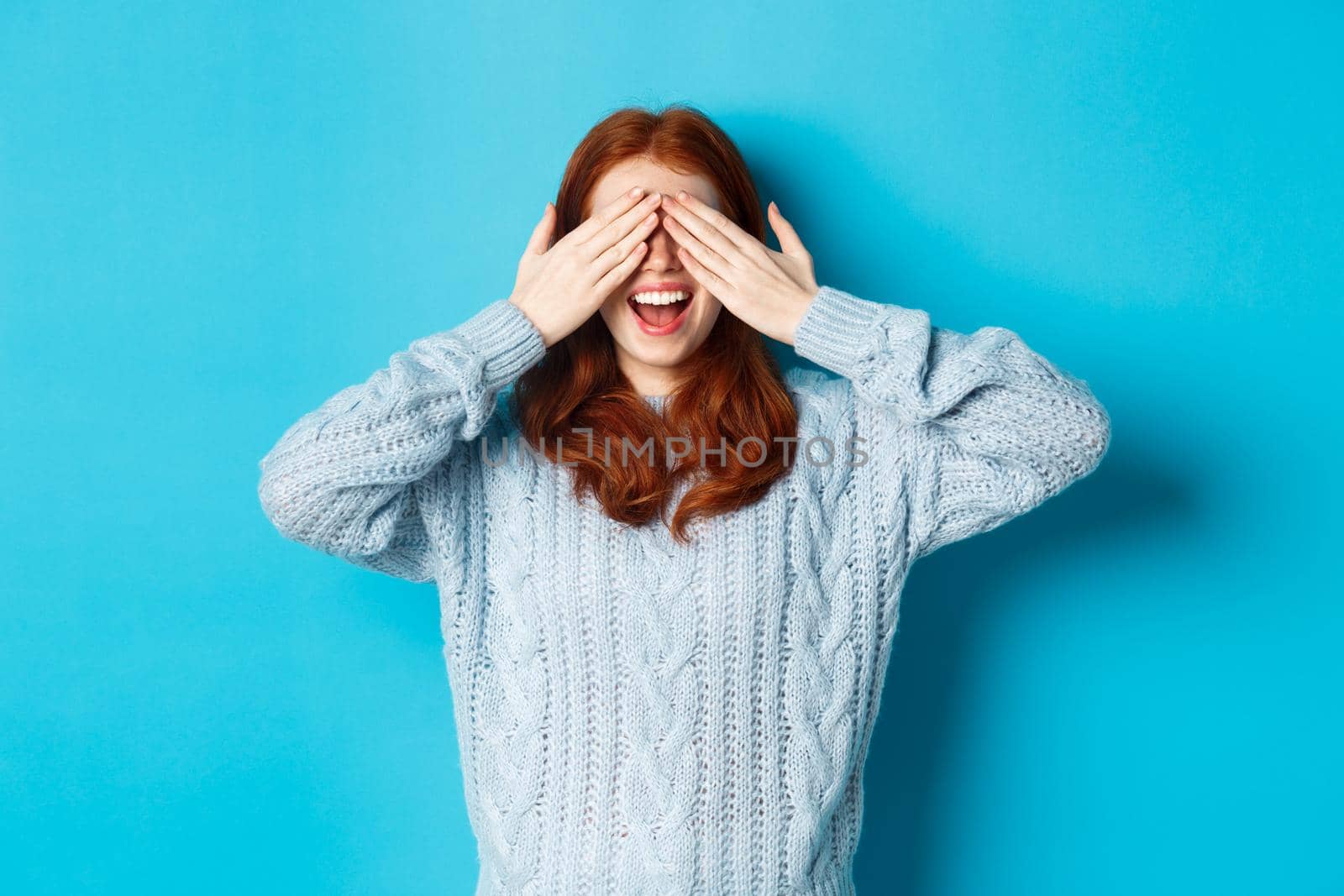 Cheerful redhead female model close eyes and waiting for christmas gift, holding hands on face and smiling amused, anticipating surprise, standing over blue background by Benzoix