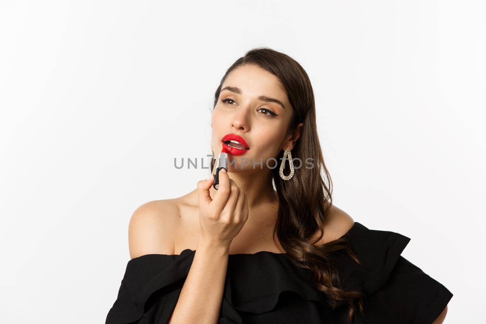 Fashion and beauty concept. Beautiful woman in black dress applying red lipstick and makeup, going on party, standing over white background.