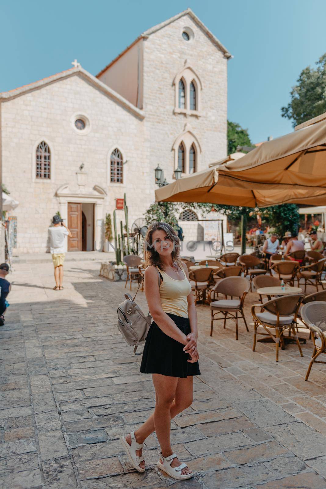 Girl Tourist Walking Through Ancient Narrow Street On A Beautiful Summer Day In MEDITERRANEAN MEDIEVAL CITY, OLD TOWN BUDVA, MONTENEGRO. Young Beautiful Cheerful Woman Walking On Old Street At Tropical Town. Pretty Girl Looking At You And Smiling by Andrii_Ko