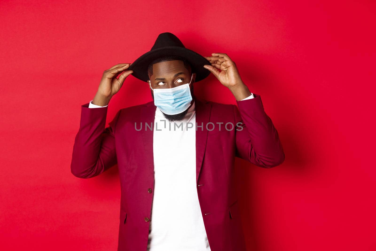 Covid-19, quarantine and holidays concept. Handsome and stylish african american man in face mask, put hat on head and looking sassy, dress-up for party, standing over red background.