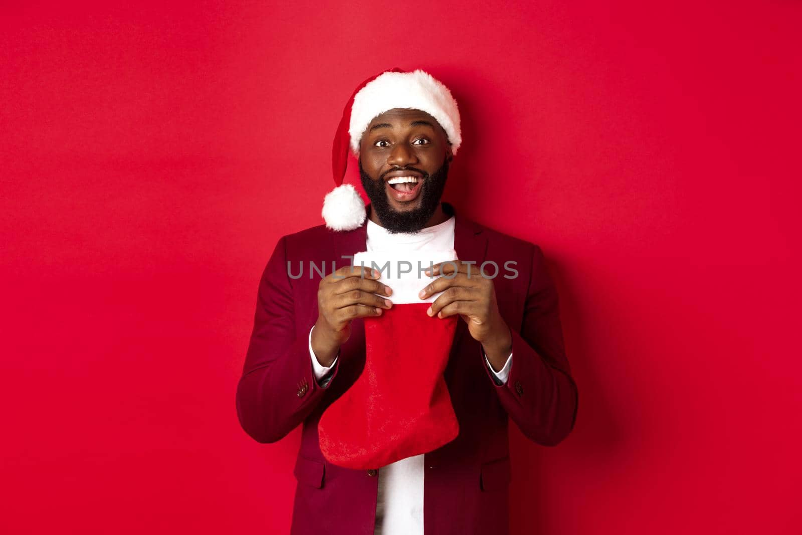 Excited black man open christmas sock with presents and sweets, smiling happy, standing in santa hat against red background by Benzoix