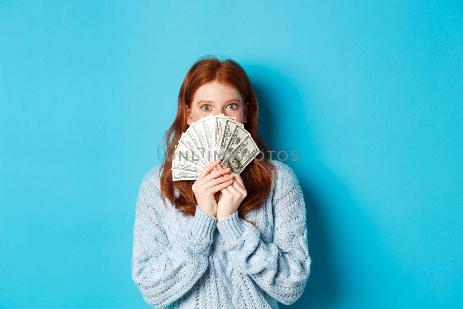 Excited redhead woman covering face with money, holding dollars and staring at camera happy, standing over blue background by Benzoix