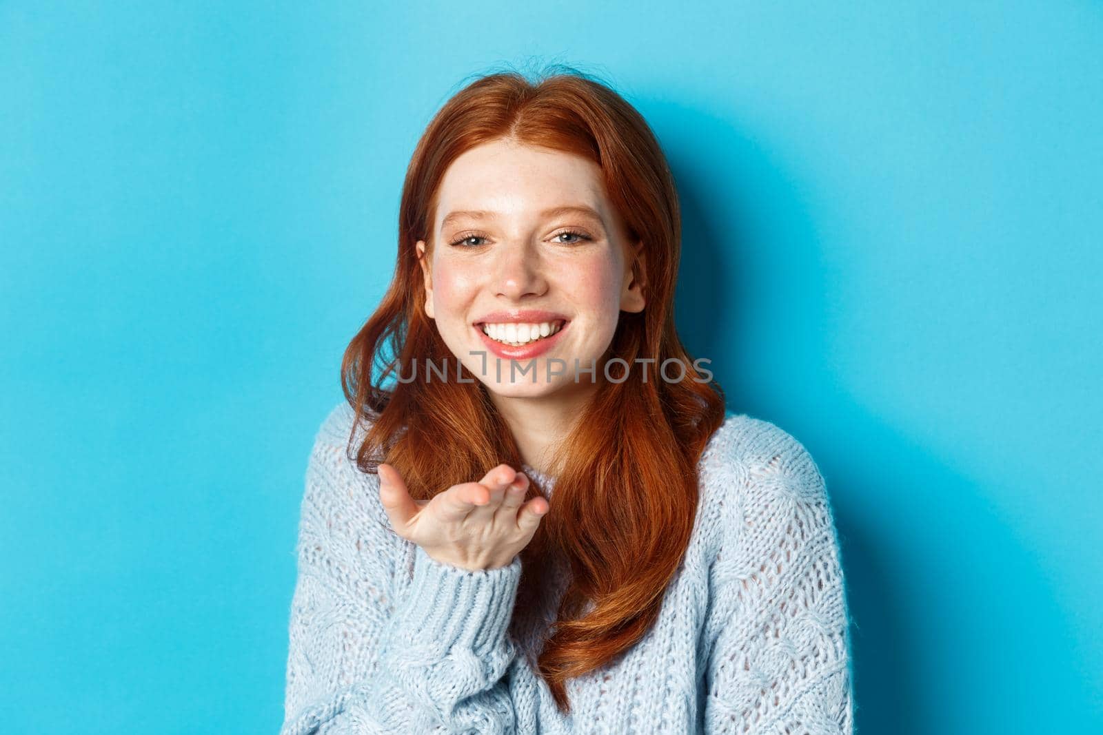 Lovely redhead female model smiling, sending air kiss at camera, standing against blue background.
