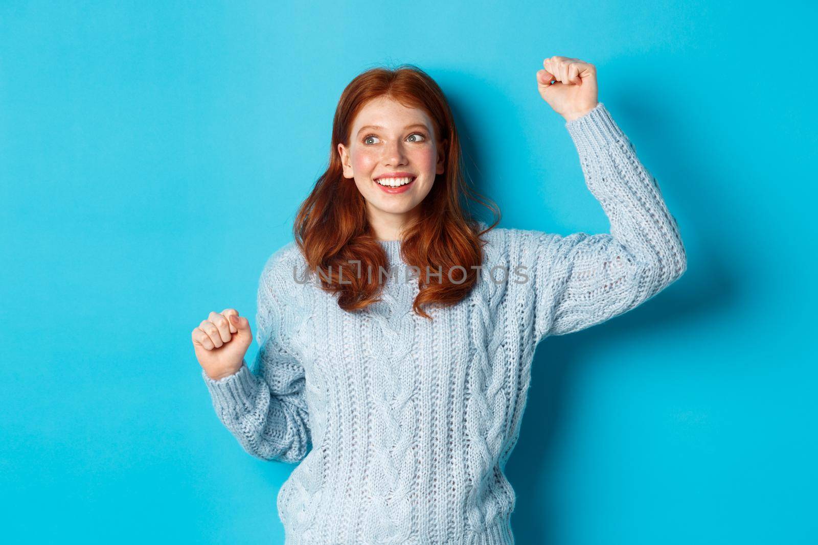 Happy redhead girl rooting for team, cheering with raised hand and smiling, celebrating victory or sucess, standing against blue background.