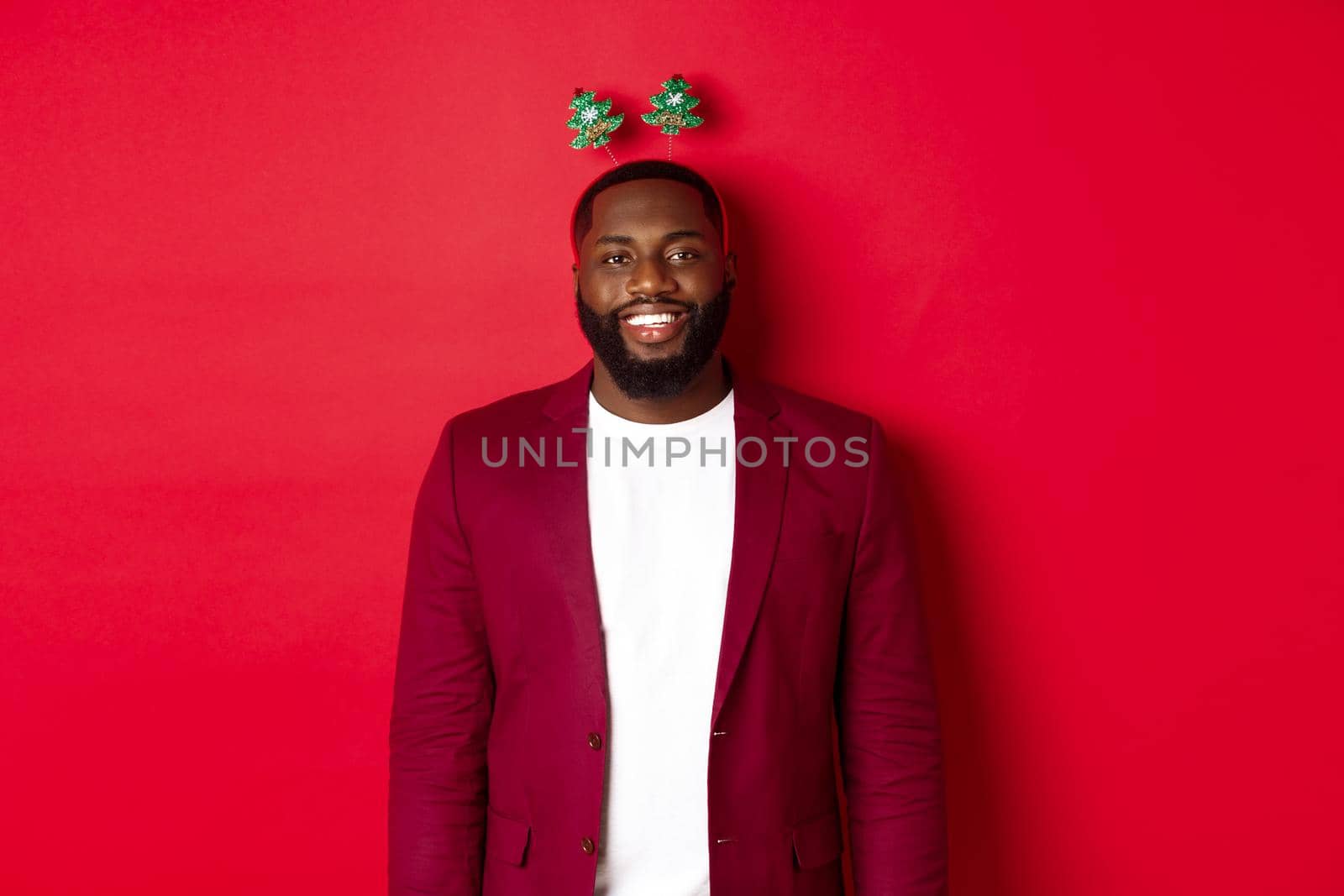 Merry Christmas. Happy african american man celebarting New Year, wearing funny party headband and smiling, standing over red background by Benzoix