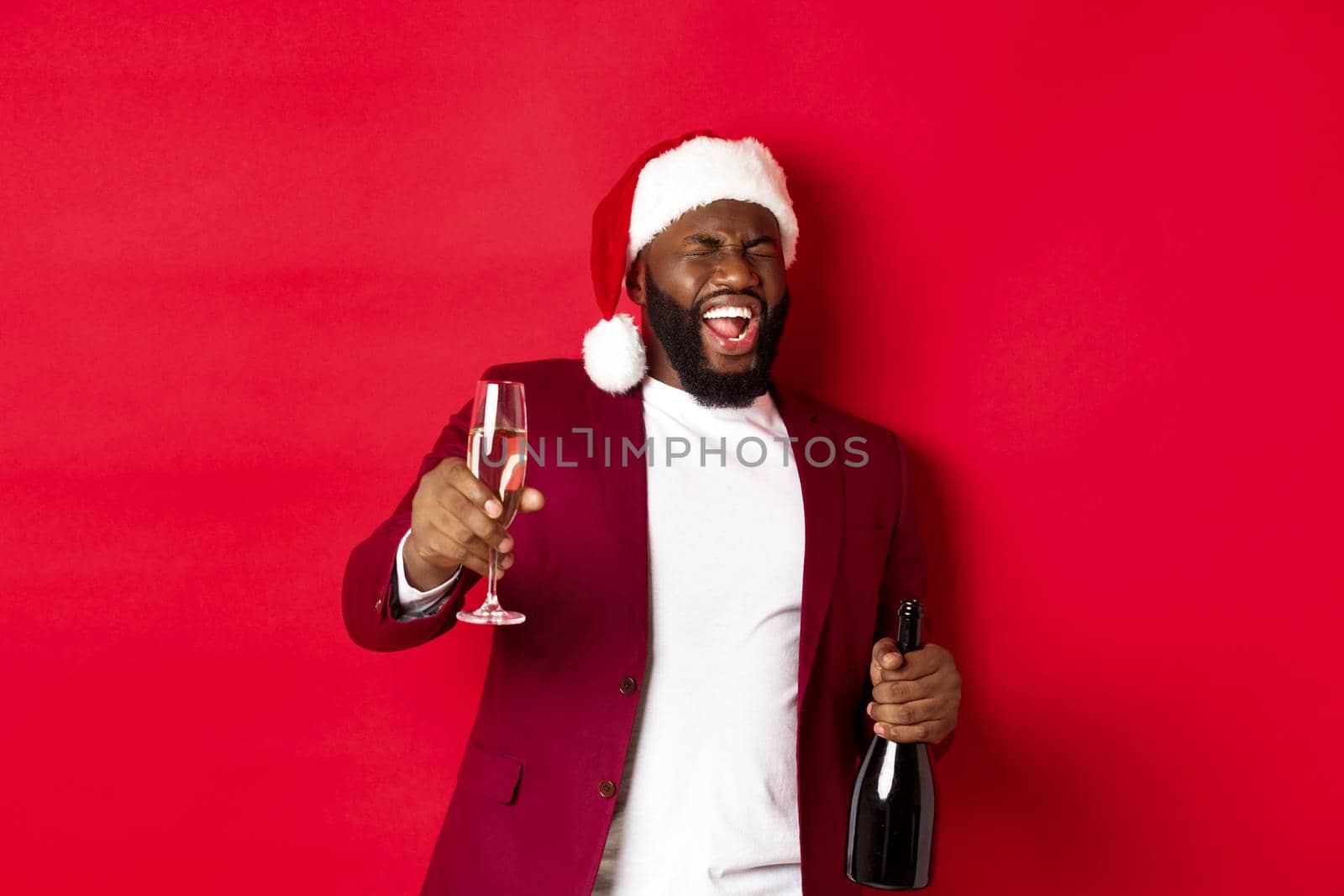 Christmas, party and holidays concept. Funny drunk Black man celebrating New Year, laughing and having fun, holding glass and bottle of champagne, red background.