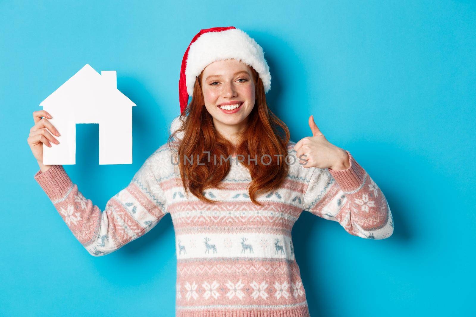 Holiday promos and real estate concept. Satisfied female model with red wavy hair, wearing santa hat and sweater, showing paper house model and thumbs-up, blue background by Benzoix