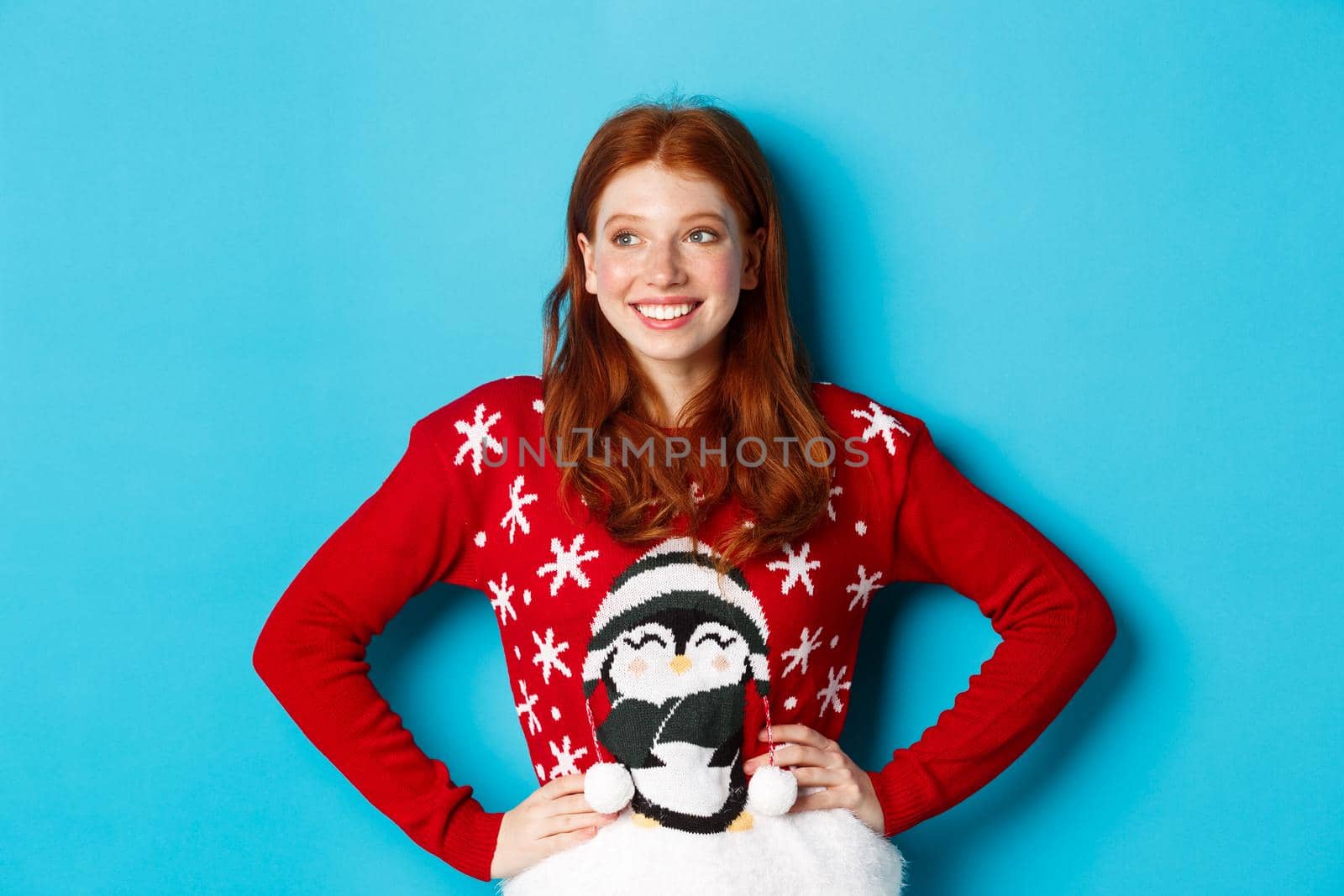 Winter holidays and Christmas Eve concept. Beautiful teenage redhead girl in xmas sweater looking left at logo, smiling pleased, holding hands on waist, blue background by Benzoix