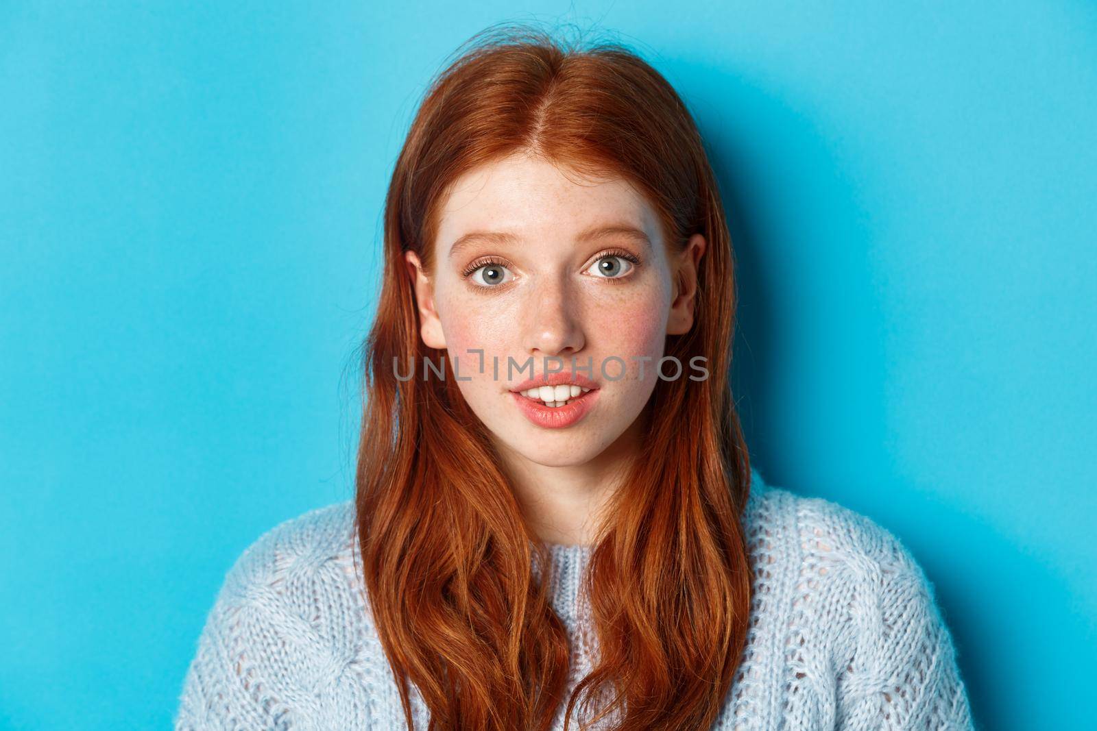 Headshot of cute redhead girl with freckles, looking hopeful and innocent at camera, standing over blue background by Benzoix