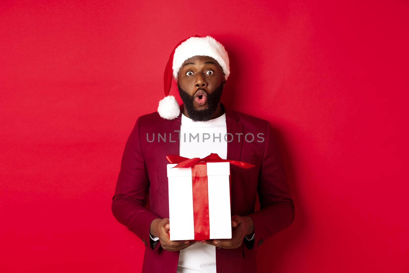 Christmas, New Year and shopping concept. Surprised african american man in santa hat saying wow, holding holiday gift and looking at camera amazed, red background.