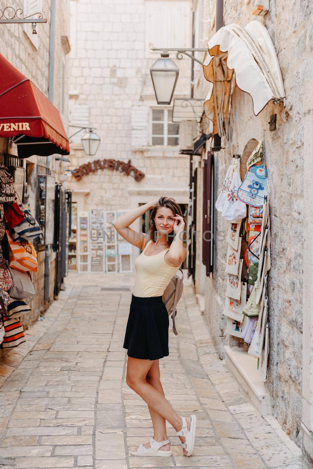 Girl Tourist Walking Through Ancient Narrow Street On A Beautiful Summer Day In MEDITERRANEAN MEDIEVAL CITY, OLD TOWN BUDVA, MONTENEGRO. Young Beautiful Cheerful Woman Walking On Old Street At Tropical Town. Pretty Girl Looking At You And Smiling by Andrii_Ko