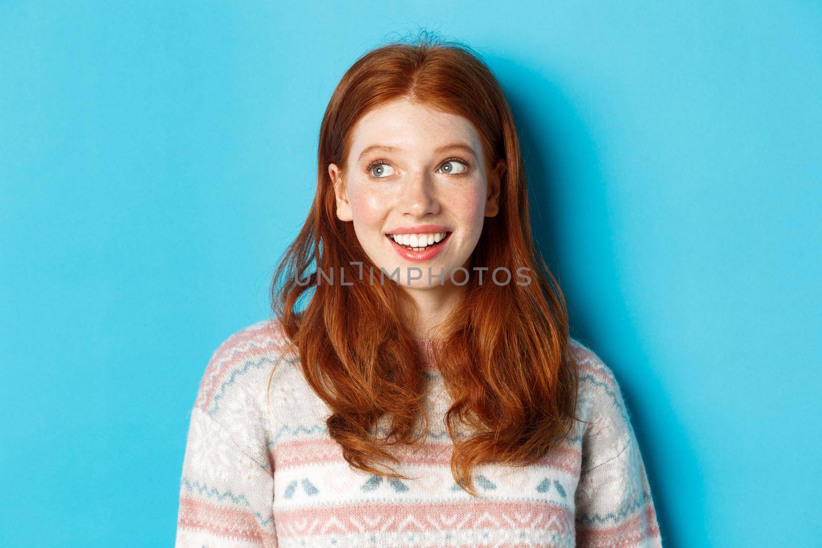 Close-up of dreamy redhead girl in winter sweater looking left, smiling and staring at promo offer, standing against blue background by Benzoix