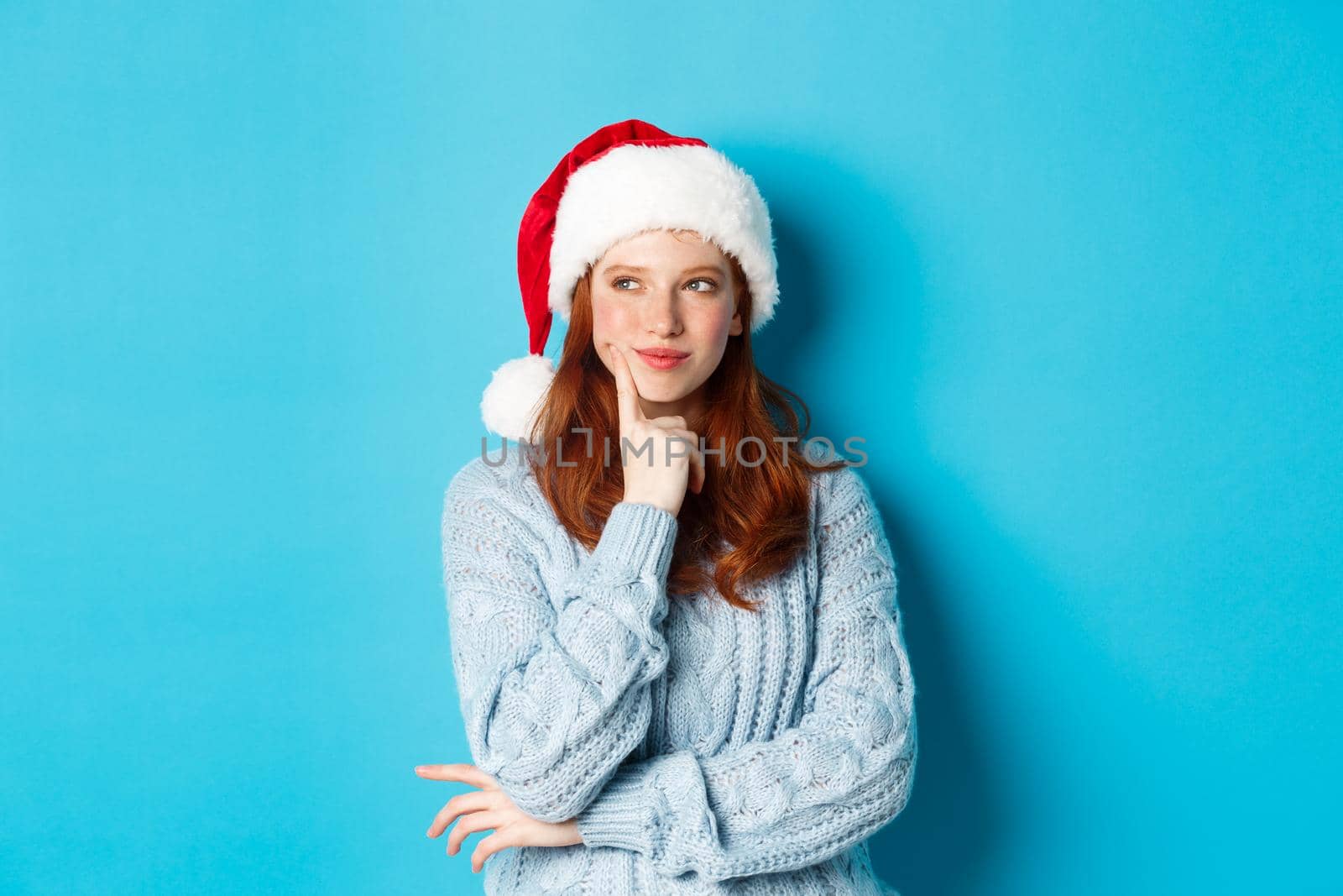 Winter holidays and Christmas Eve concept. Silly redhead girl with freckles, wearing santa hat and thinking, planning New Year celebration, standing over blue background.