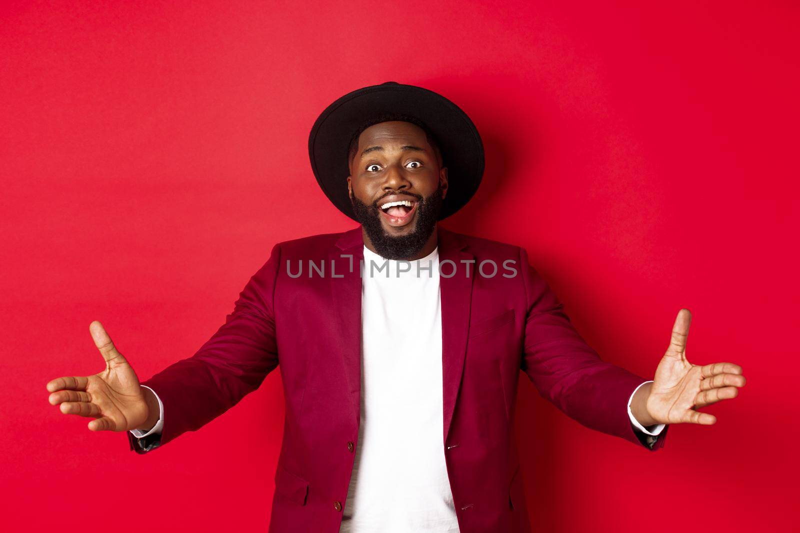 Fashion and party concept. Pleased handsome Black man spread hands in welcome gesture, reaching to receive something, looking amazed, standing over red background by Benzoix