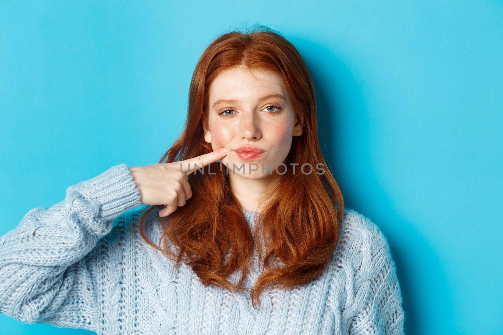 Close-up of sassy teen girl with red hair, pointing at her cheek and staring at camera, standing against blue background by Benzoix