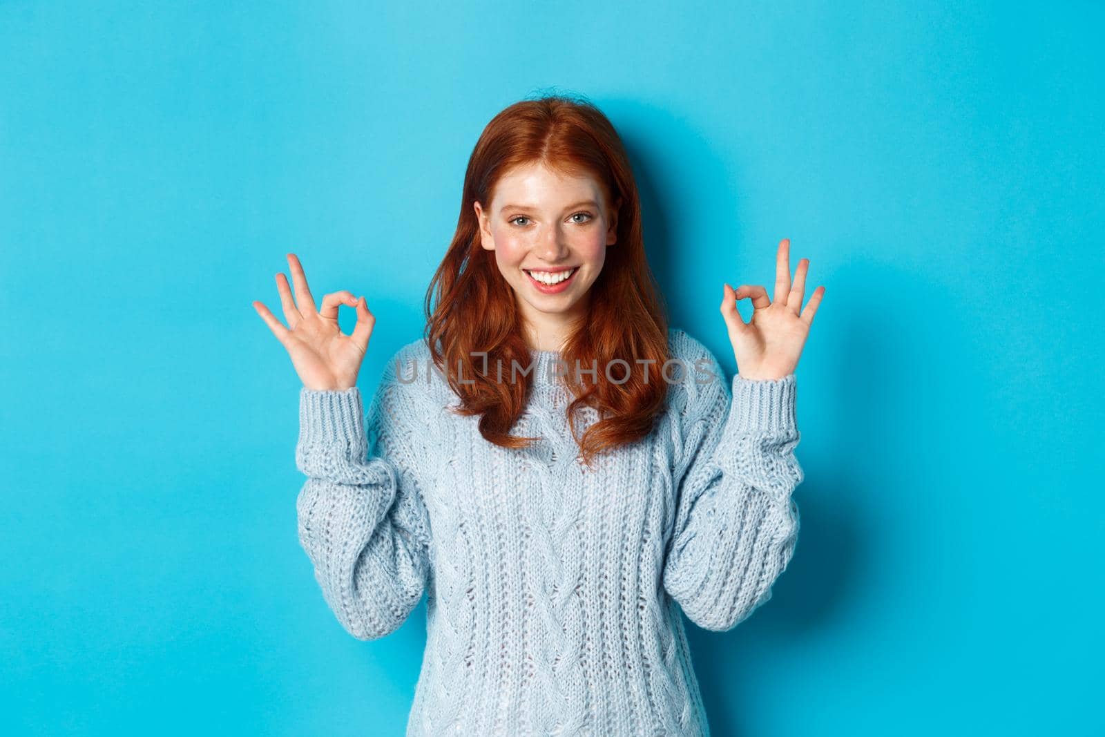 Confident redhead girl showing okay signs, praising choice, approve and agree, like something, standing over blue background by Benzoix