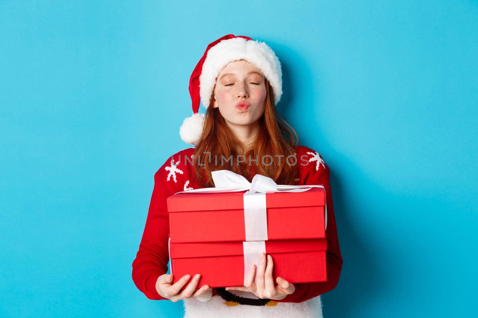 Happy holidays and Christmas concept. Cute redhead girl holding presents and pucker lips for kiss, wearing santa hat and funny sweater, blue background by Benzoix