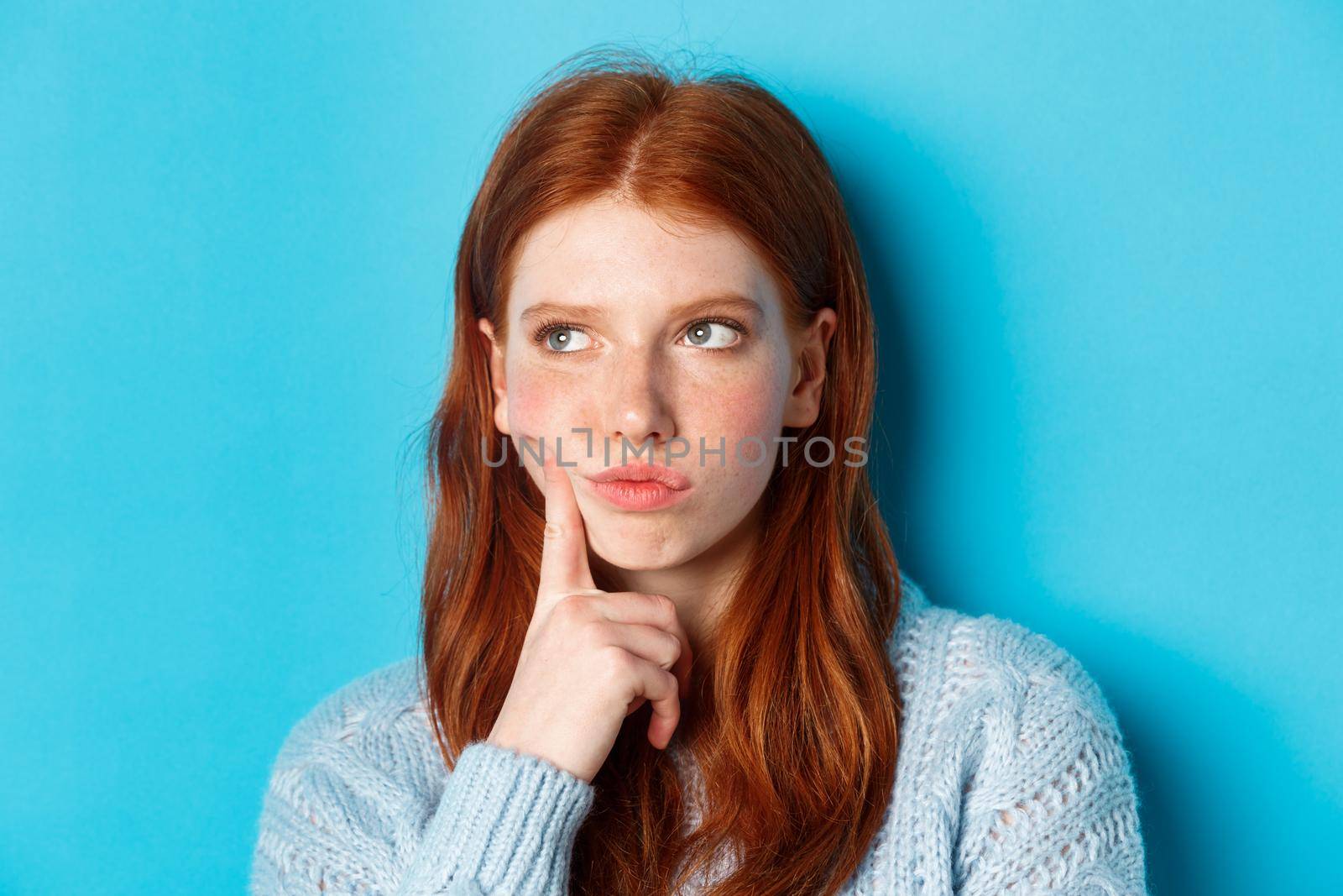 Headshot of troubled teenage girl thinking, looking bothered and frowning, standing against blue background.