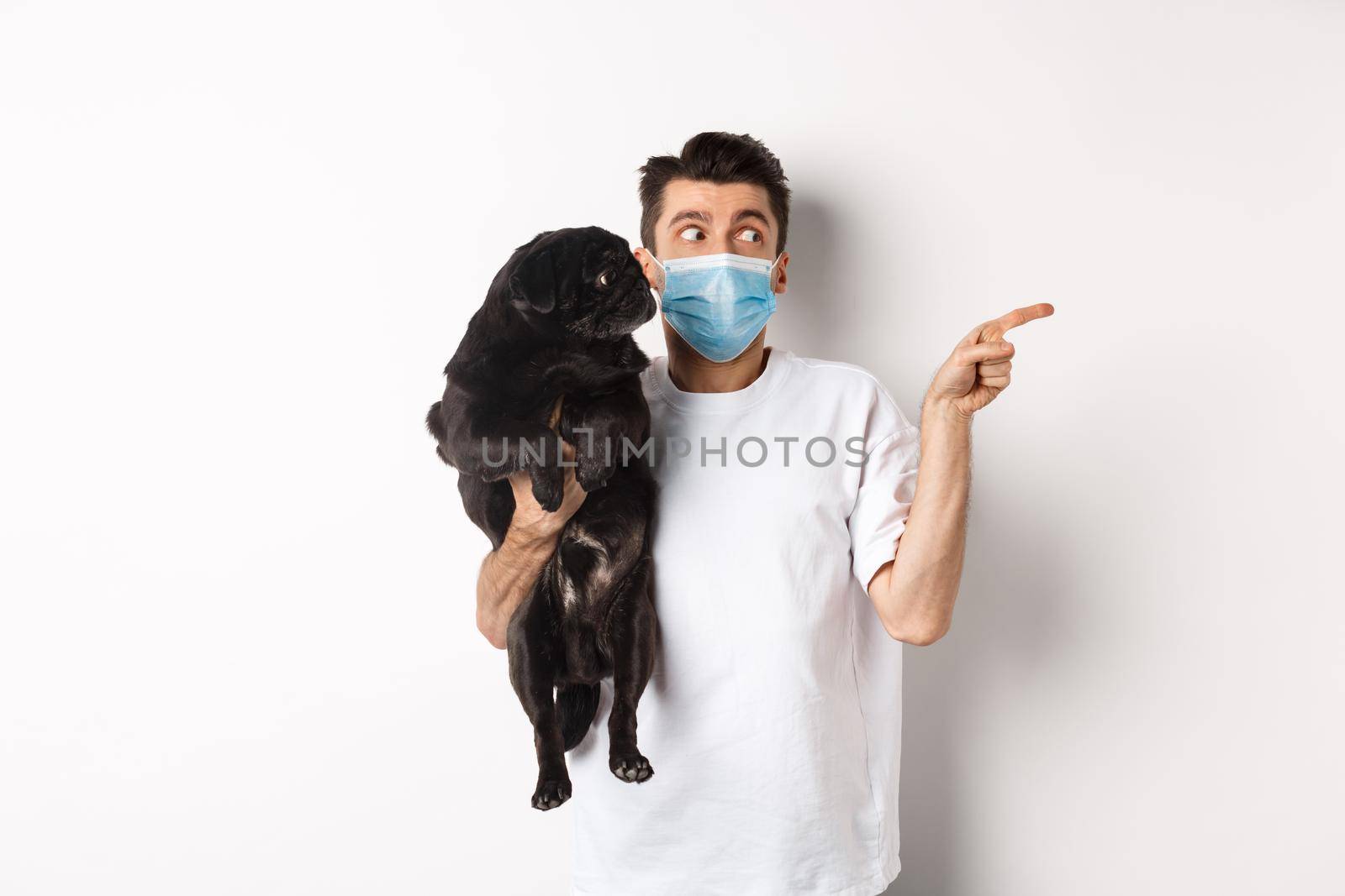 Covid-19, animals and quarantine concept. Young man in face mask holding cute black pug, dog and pet owner looking right at copy space, standing over white background.
