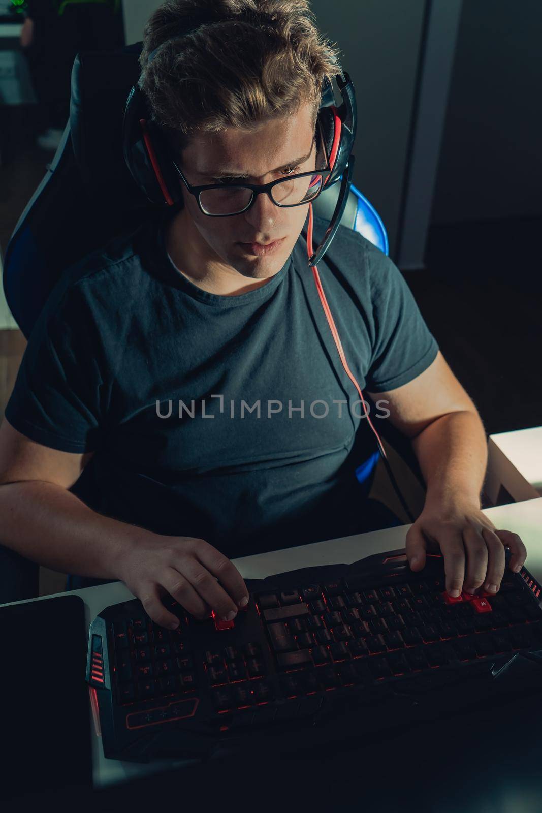 Young man with short blond hair and glasses, cheerful and happy, dressed in black shirt, playing video games, playing on his laptop in the dark illuminated by the screen. Dim light from computer monitor, dark room, desk with keyboard, computer screen and coloured lights. Vertical