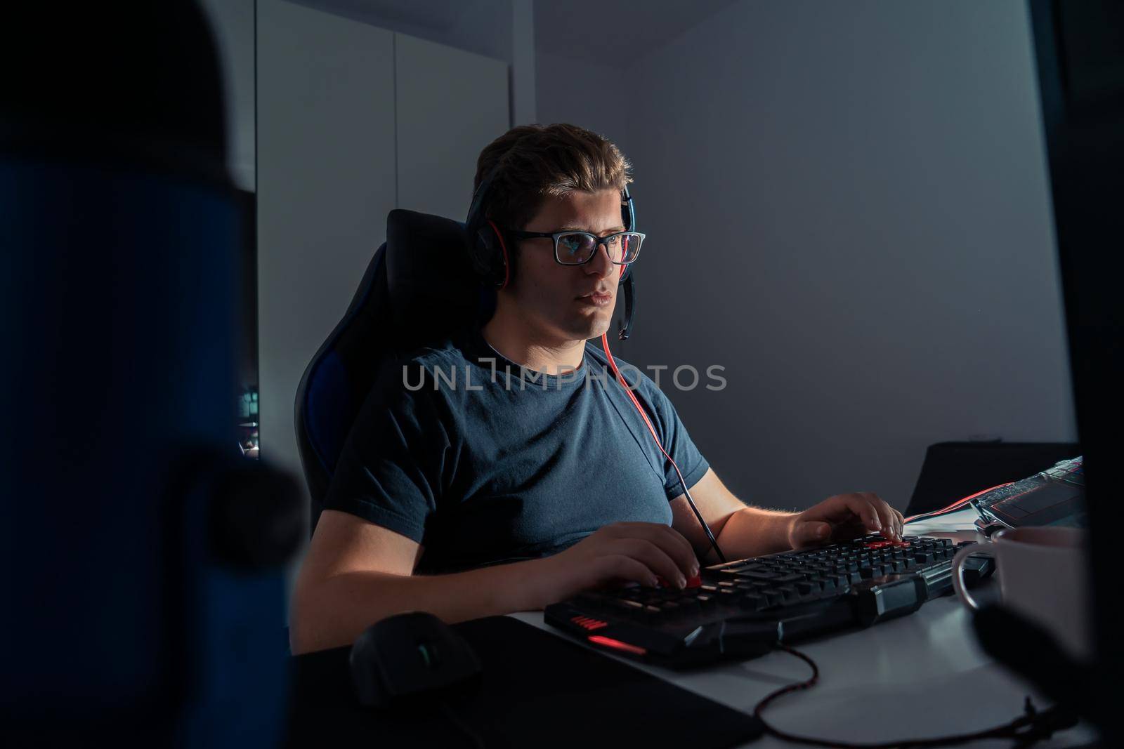 Short blond-haired young man with glasses, cheerful and happy, dressed in black shirt, fond of video games, playing on his laptop computer at night. Young video game player. Dim light from computer monitor, dark room, desk with keyboard, computer screen and coloured lights. Horizontal