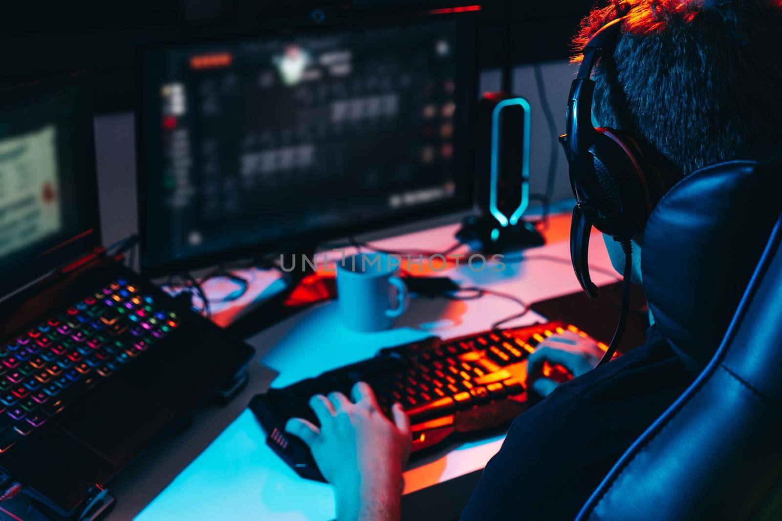 Young man with short blond hair and glasses, and headphones, dressed in black shirt, addicted to video games playing on computer with headphones. Young gamer illuminated with blue and red coloured lights. Dim light from computer monitor, dark room, desk with keyboard, computer screen and coloured lights. Horizontal.