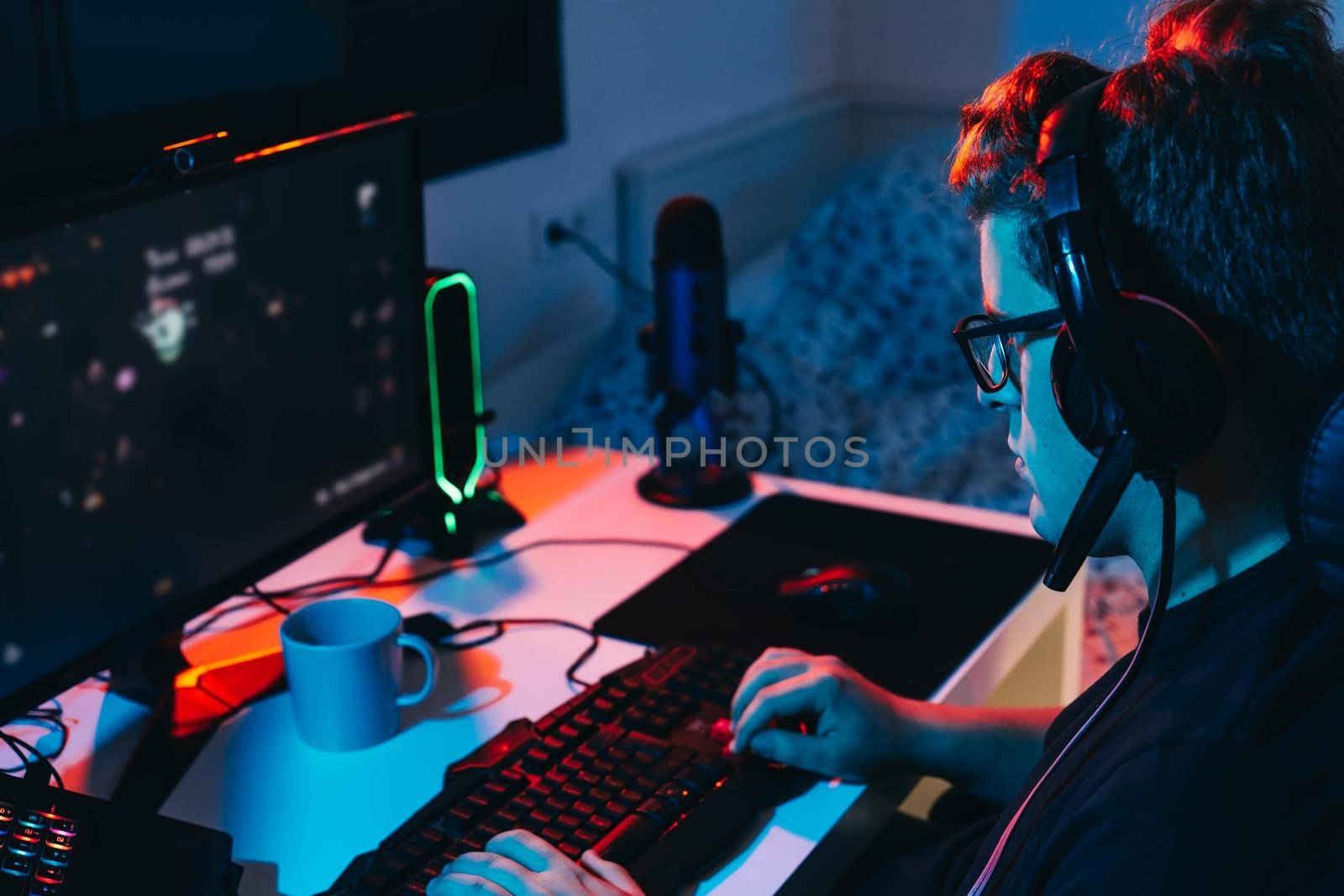 Young man with short blond hair, wearing glasses and headphones, dressed in black shirt, addicted to online video games, playing on a computer with headphones. Young gamer illuminated with blue and red coloured lights. Dim light from computer monitor, dark room, desk with keyboard, computer screen and coloured lights. Horizontal.