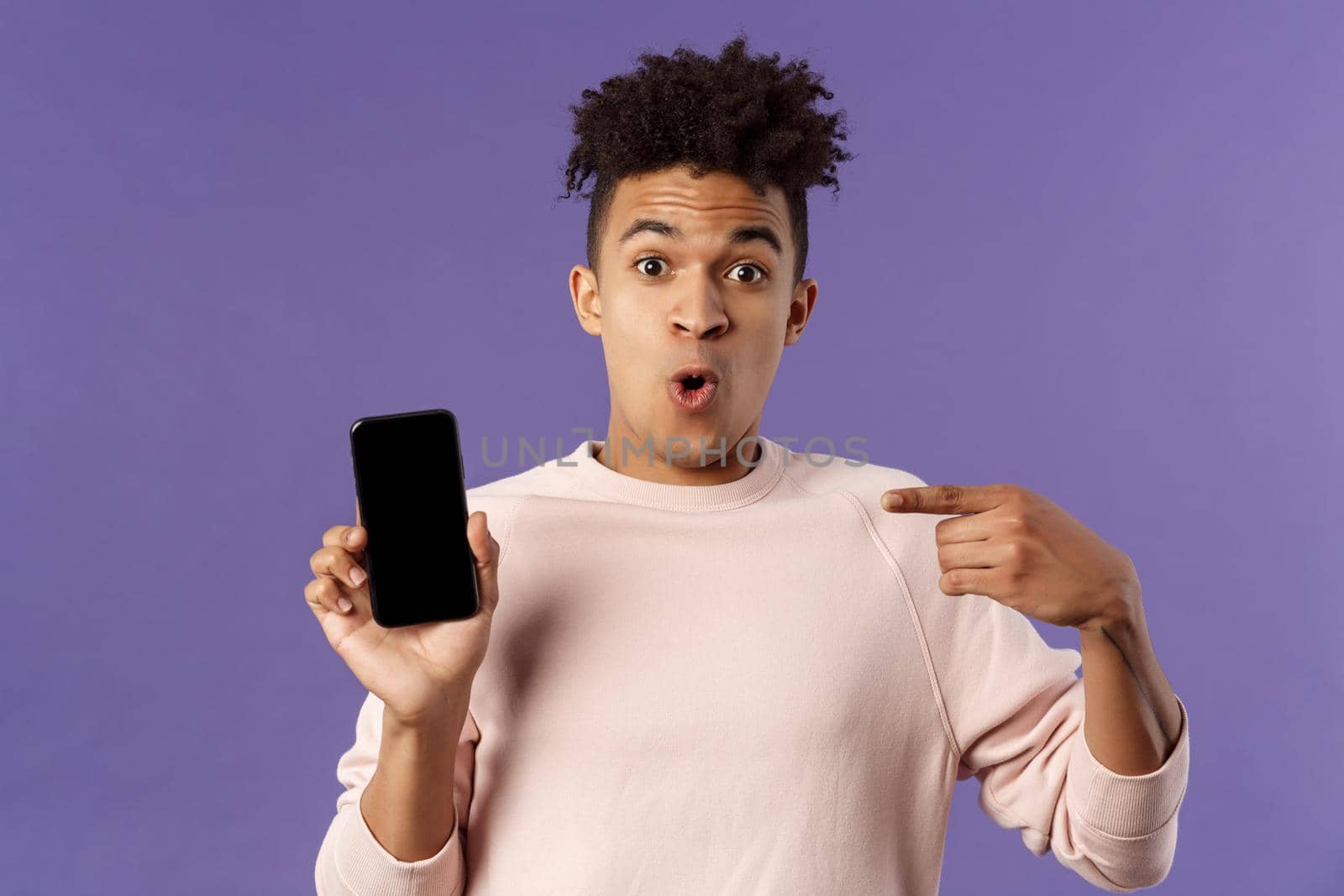 Close-up portrait of excited, cheerful young hispanic male geek talking about his device, new gadget or application, holding mobile phone, pointing at smartphone with amused face by Benzoix
