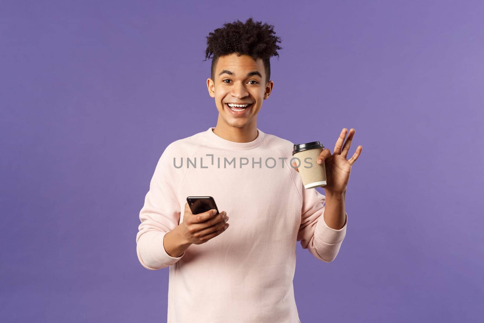 Portrait of cheerful young male student with cup of coffee and mobile phone, waving hand to say hi, smiling friendly as greeting person, meet classmate near university campus, purple background by Benzoix