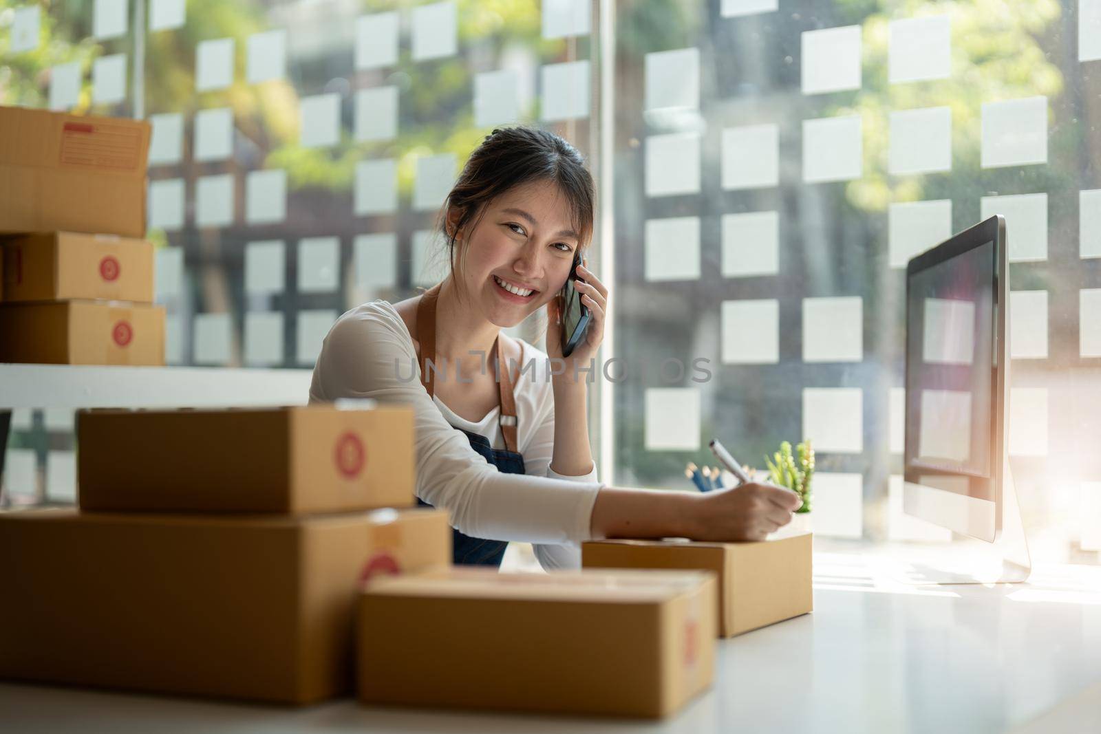 Young Asian small business owner working at home office, taking note on purchase orders. Online marketing packaging delivery, startup SME entrepreneur or freelance woman concept.