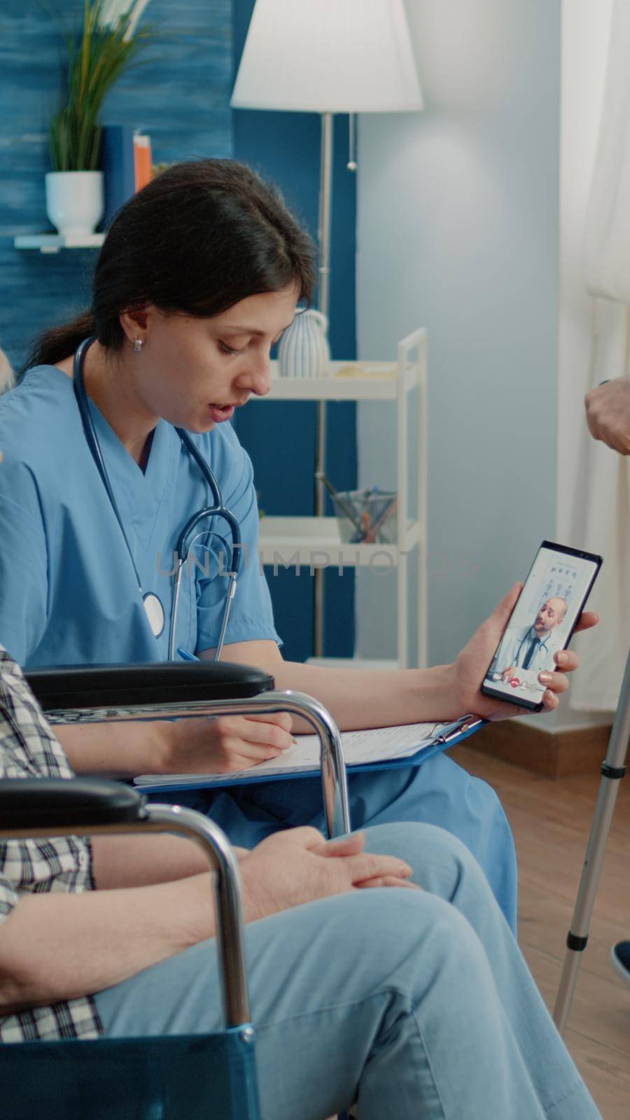 Retired woman and nurse talking to doctor on video call for telemedicine and telehealth in nursing home. Medical assistant holding smartphone, helping disabled patient in weelchair