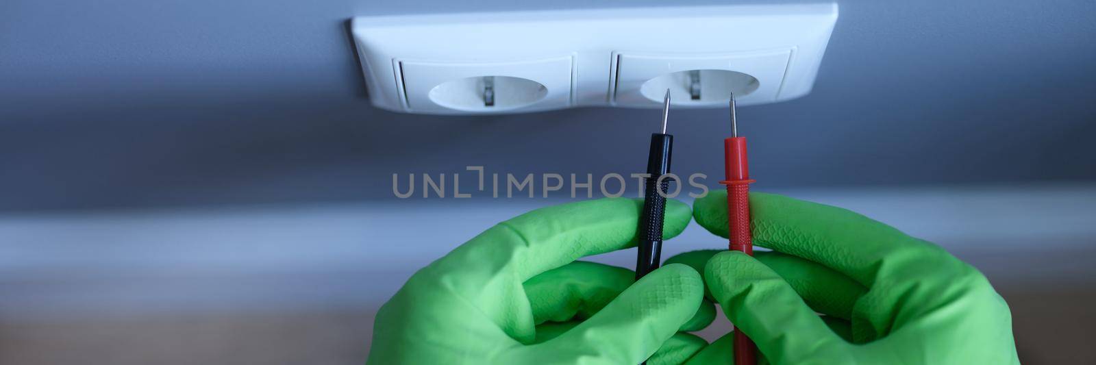 Craftsman in rubber gloves holding tester near electrical outlet at home closeup. Safety when working with electricity concept