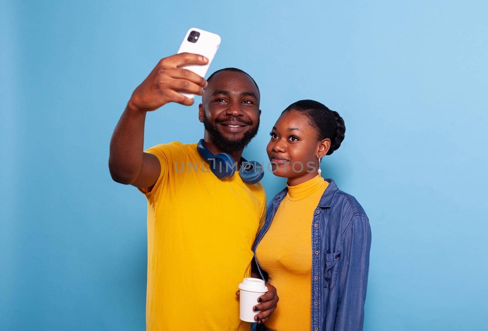Happy couple taking selfies on smartphone in studio, making memories together. Modern boyfriend and girlfriend taking pictures on mobile phone, embracing and using technology over background.