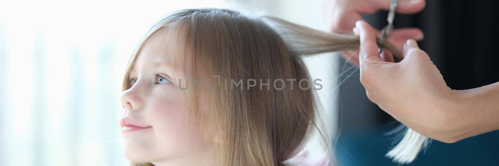 Hairdresser cutting hair of little girl in beauty salon. Hairdressing services for children concept