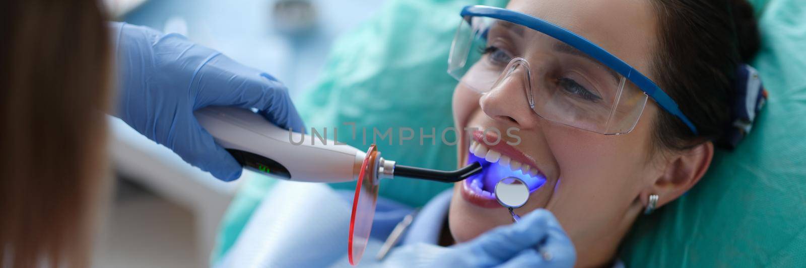 Young woman dentist in protective gloves checking dental seal, fixing photopolymer lamp during stomatology procedure by kuprevich