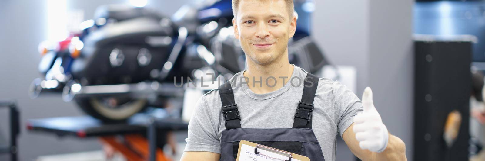 Portrait of young car mechanic holding clipboard and thumb up in car service by kuprevich