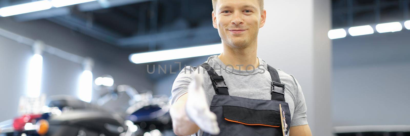 Young smiling master man in uniform stretches his arms forward in car service. High-quality service of motorcycles and cars concept