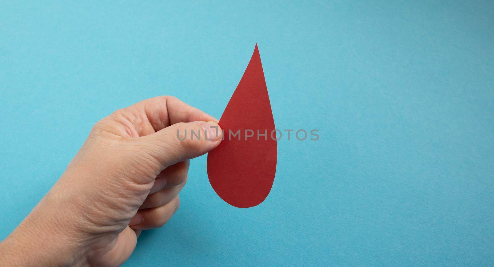 The concept of the World Blood Donor and Hemophilia Day. Red paper drop of blood in the hand on a blue background.
