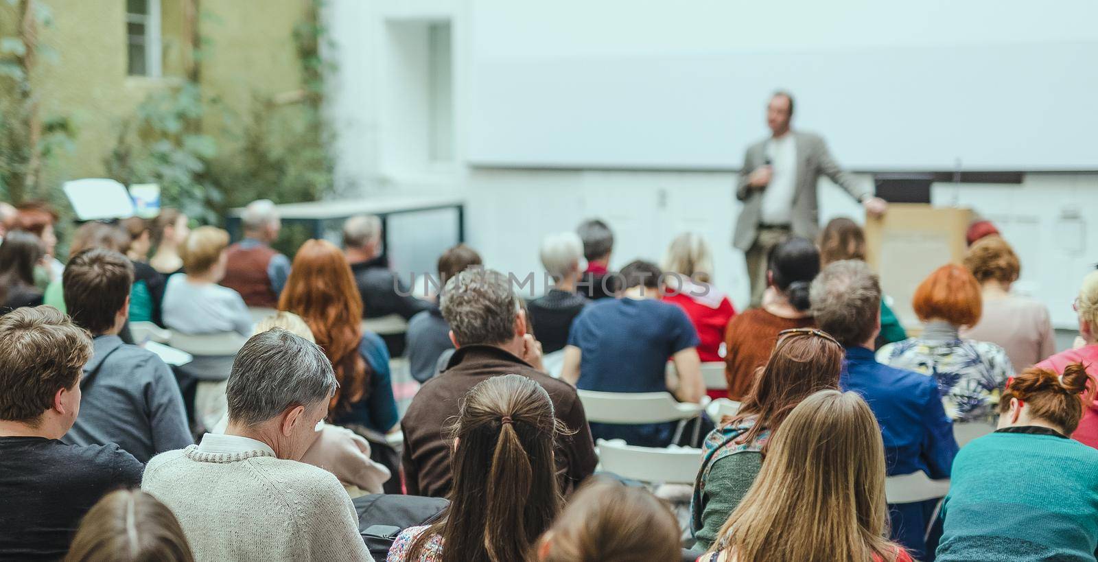 Male speaker giving presentation in lecture hall at university workshop. Audience in conference hall. Rear view of unrecognized participant. Scientific conference event. Copy space on whitescreen.