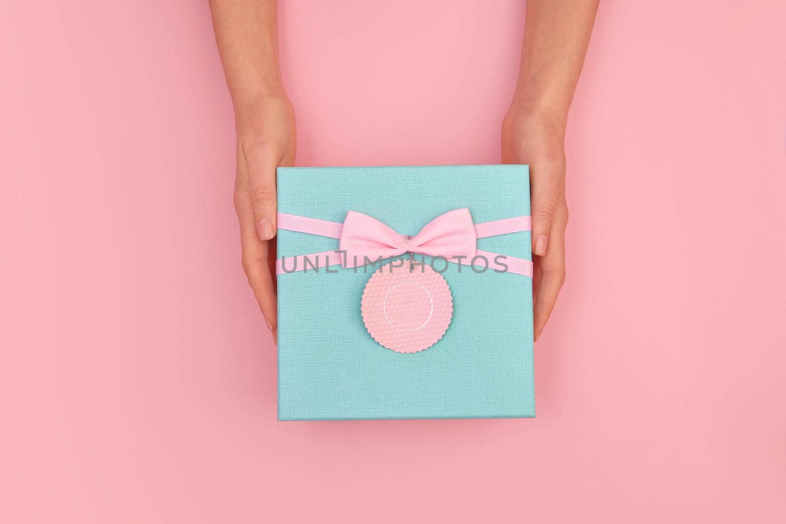 Hands give turquoise gift box with a card and a bow on isolated pastel pink background top view
