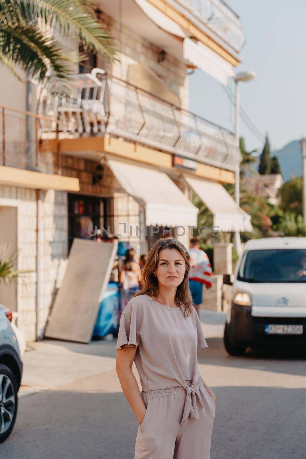 Girl Tourist Walking Through Ancient Narrow Street On A Beautiful Summer Day In MEDITERRANEAN MEDIEVAL CITY, MONTENEGRO. Young Beautiful Cheerful Woman Walking On Old Street At Tropical Town. Pretty Girl Looking At You And Smiling by Andrii_Ko