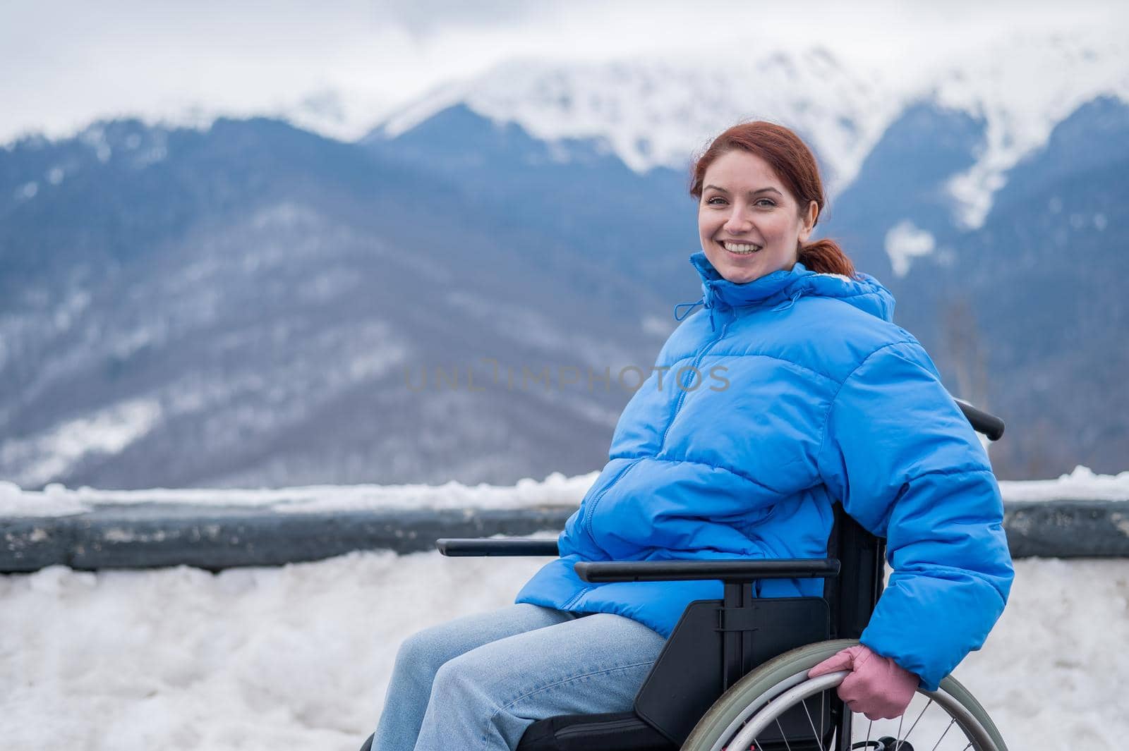 A happy woman dressed in a blue coat sits in a wheelchair on a point view and looking at the snow-capped mountains