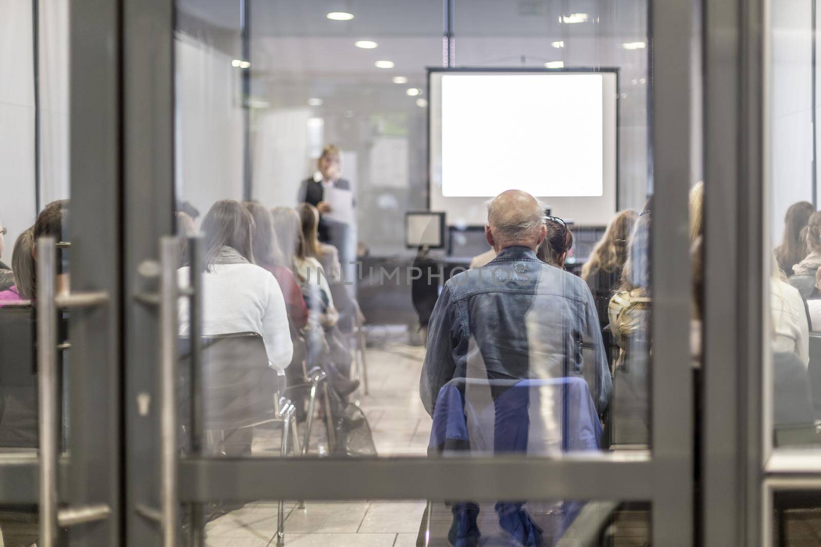 Business and entrepreneurship symposium. Female speaker giving a talk at business meeting. Audience in conference hall. Rear view of unrecognized participant in audience. Copy space on whitescreen.