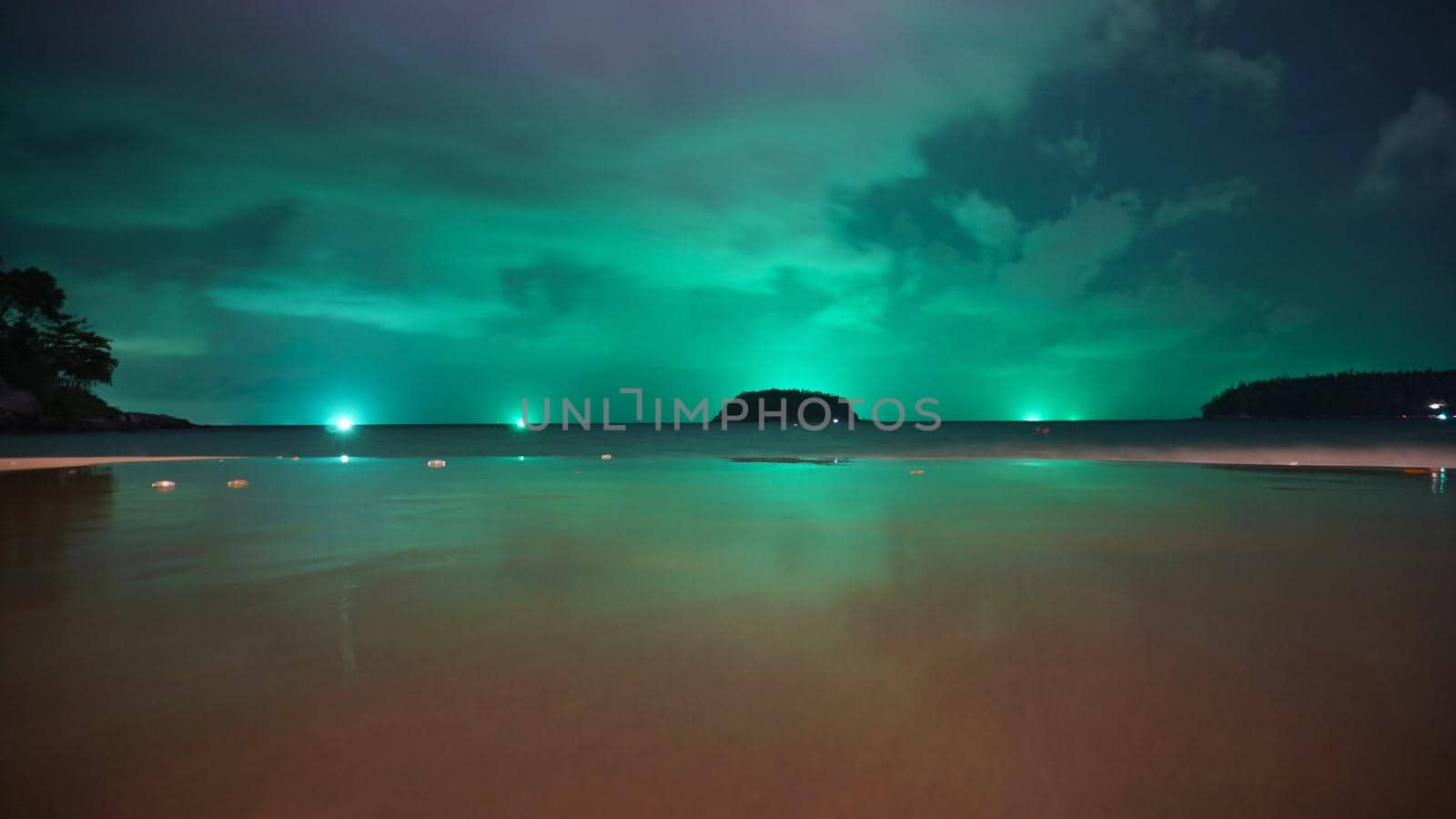 Unusual green illumination of the sky over the island. Clouds of green-purple color are reflected in the ocean, in the water and on wet sand. In distance there is an island, palm trees and lanterns