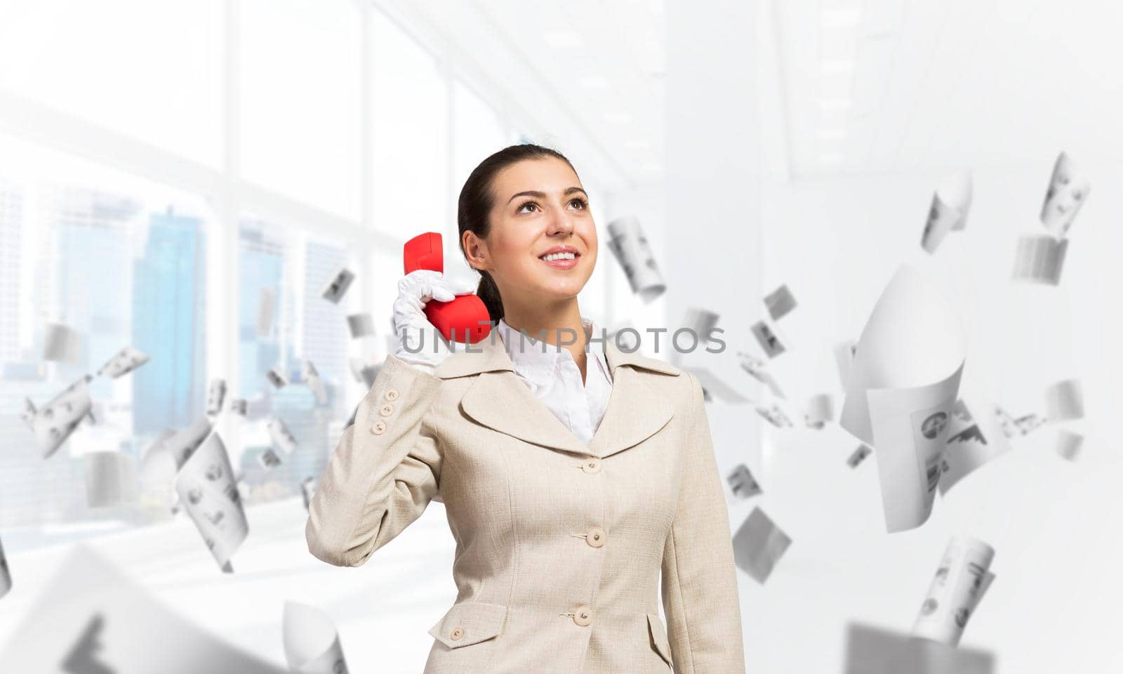Smiling young woman holding retro red phone in office with flying paper documents. Call center operator in business suit with telephone. Hotline telemarketing. Business assistance and consultation.