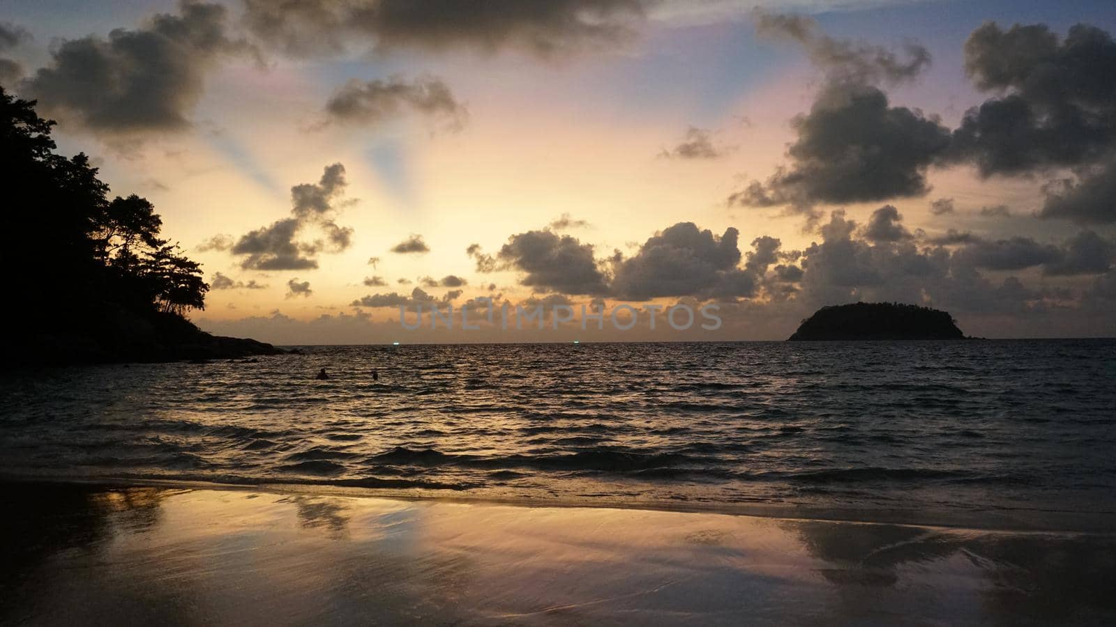 View of the beach at sunset, sea and clouds. Large clouds float across the sky. A lonely island can be seen in the distance. People relax, swim, take pictures and enjoy the sunset. Kata Beach, Phuket